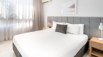 Ett modernt hotellrum med en stor säng, en grå sänggavel, ett nattduksbord med en lampa och en inramad tavla på väggen.