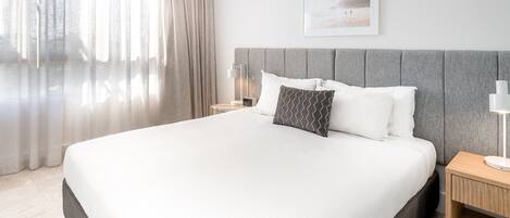 A modern hotel room with a large bed, a grey headboard, a bedside table with a lamp, and a framed picture on the wall.