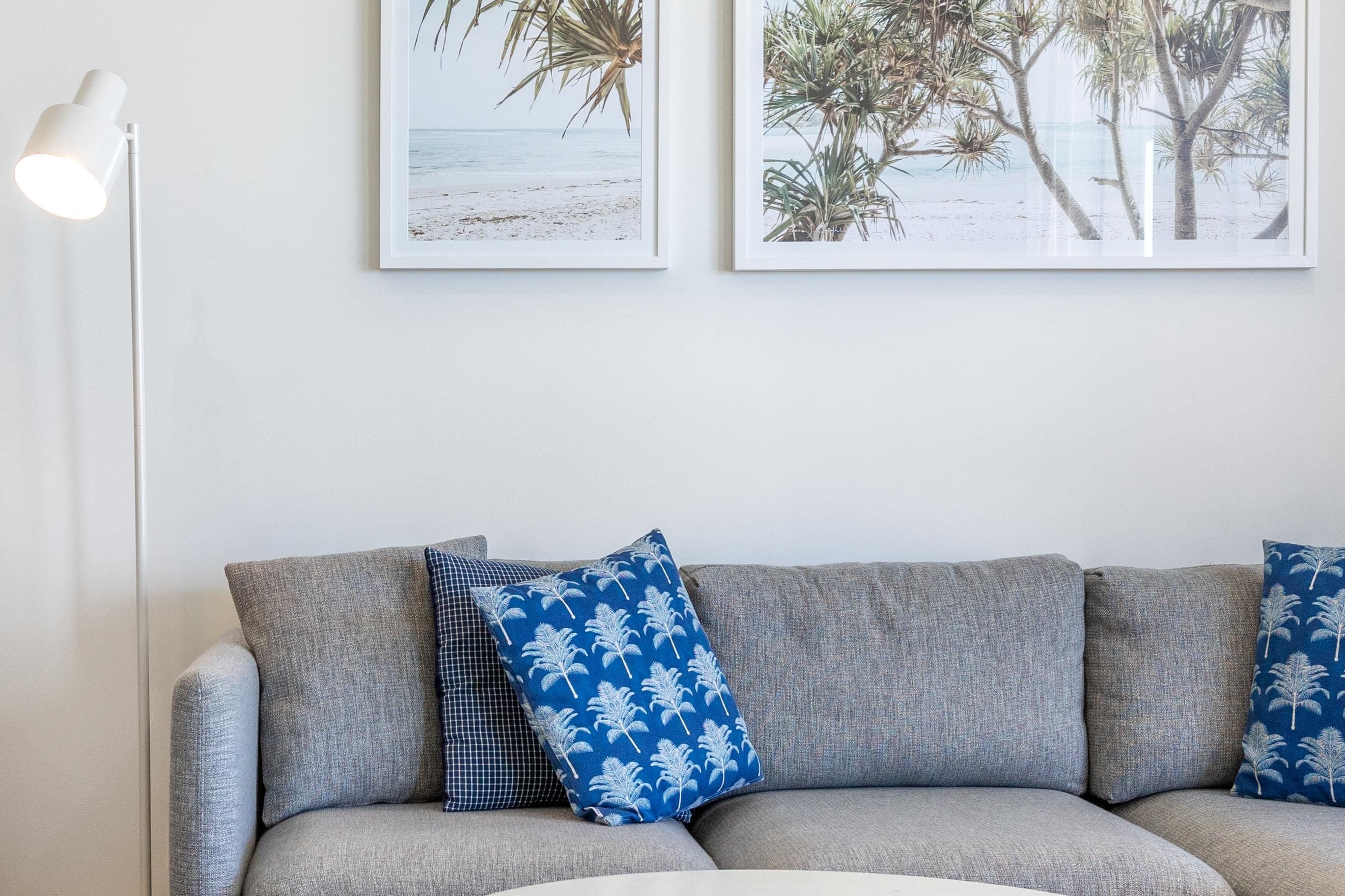 A grey sofa with patterned cushions, a white floor lamp, and two framed beach-themed pictures on the wall.