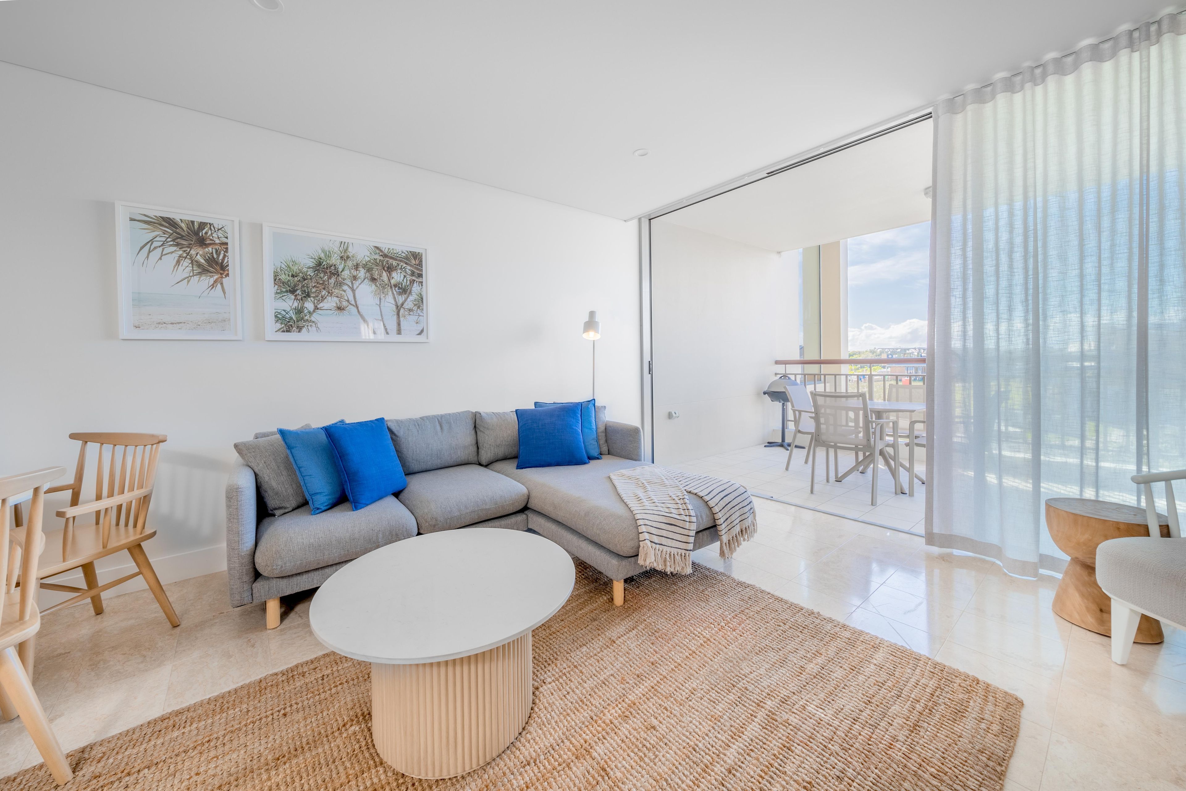 A modern living room with a grey sectional sofa, blue cushions, a round coffee table, and a large window with sheer curtains.