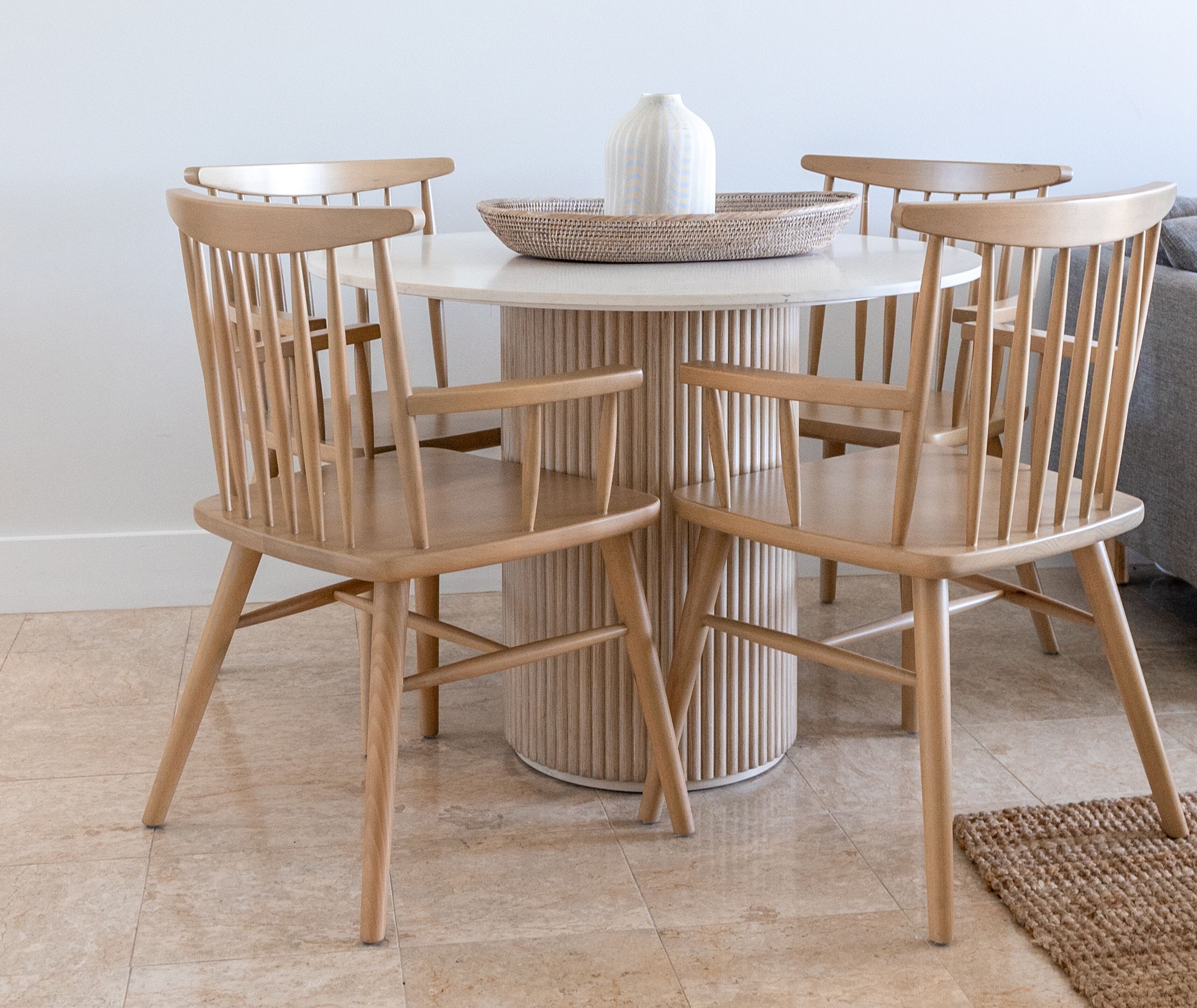 A round wooden table with four chairs, a woven basket, and a tall vase.
