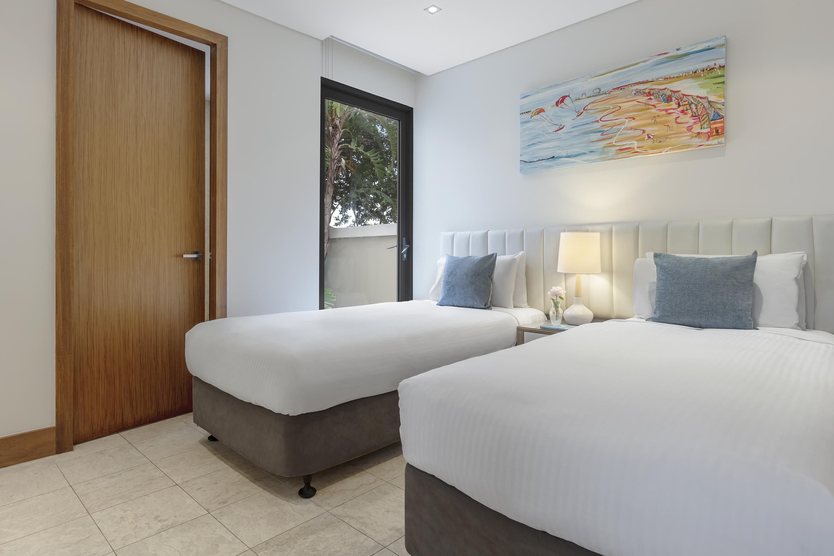A hotel room with two beds, a wooden door, a window with a view of trees, and a painting on the wall.