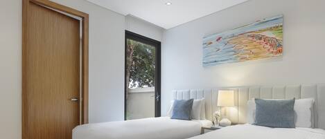A hotel room with two beds, a wooden door, a window with a view of trees, and a painting on the wall.