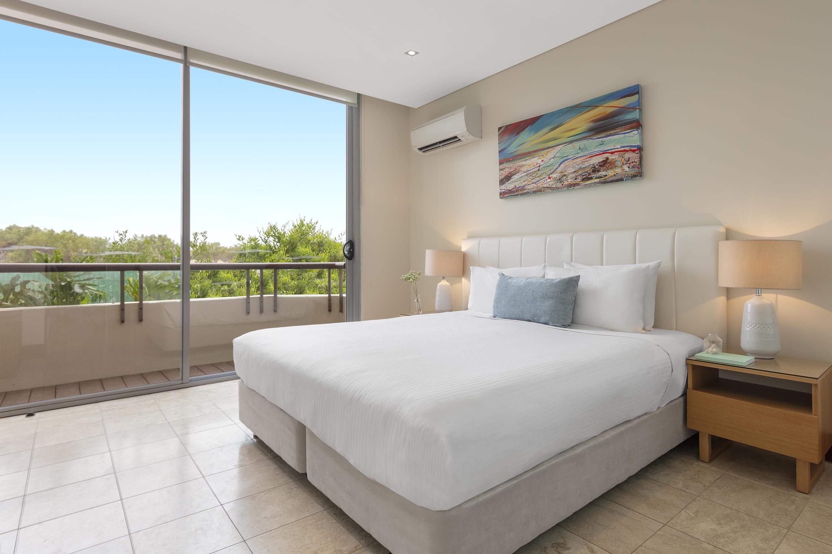 A hotel room with a large bed, two bedside lamps, a nightstand, a painting, and a balcony with a view of greenery.