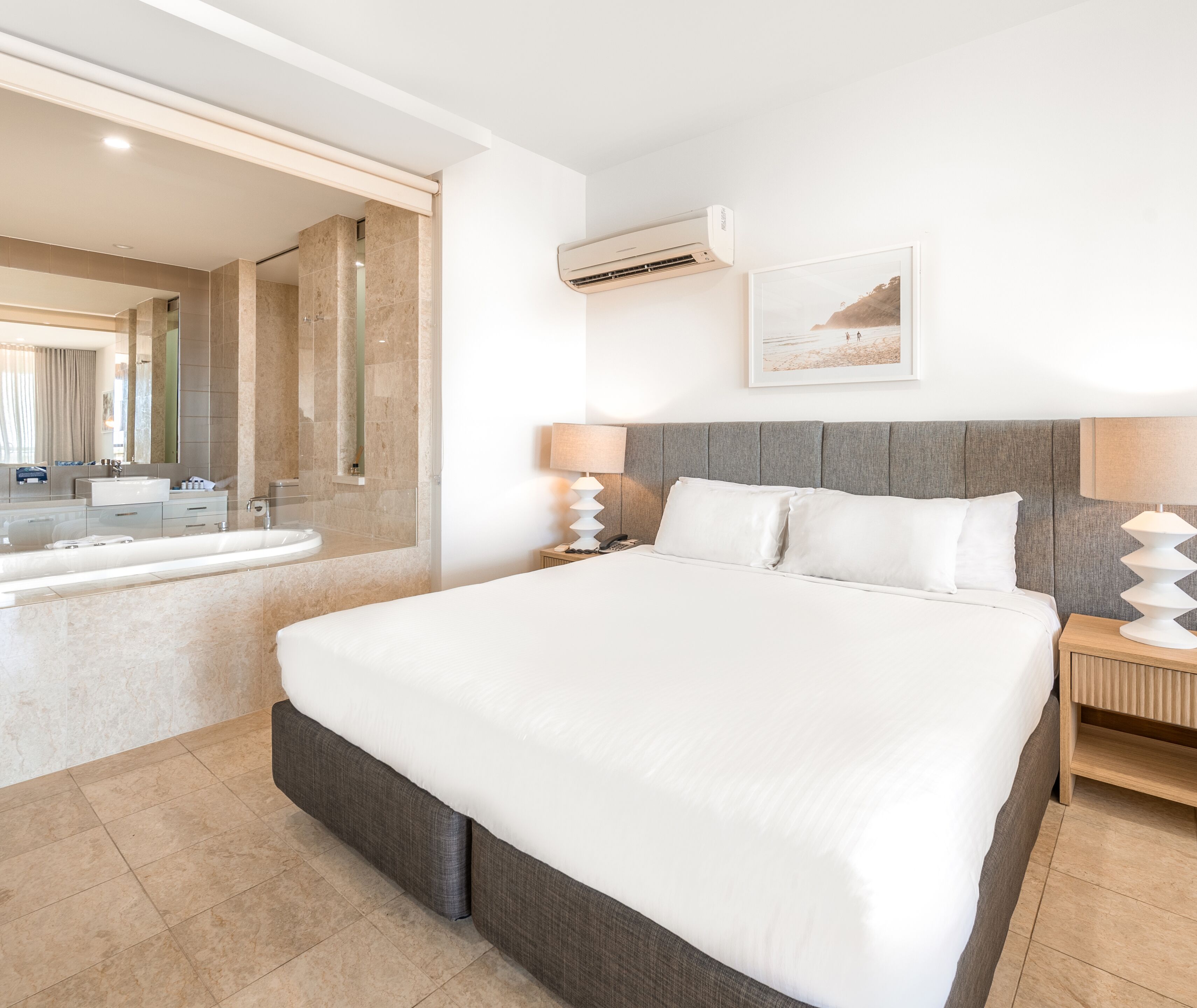 Quarto de hotel moderno com uma cama grande, banheiro com banheira e um espelho amplo.
