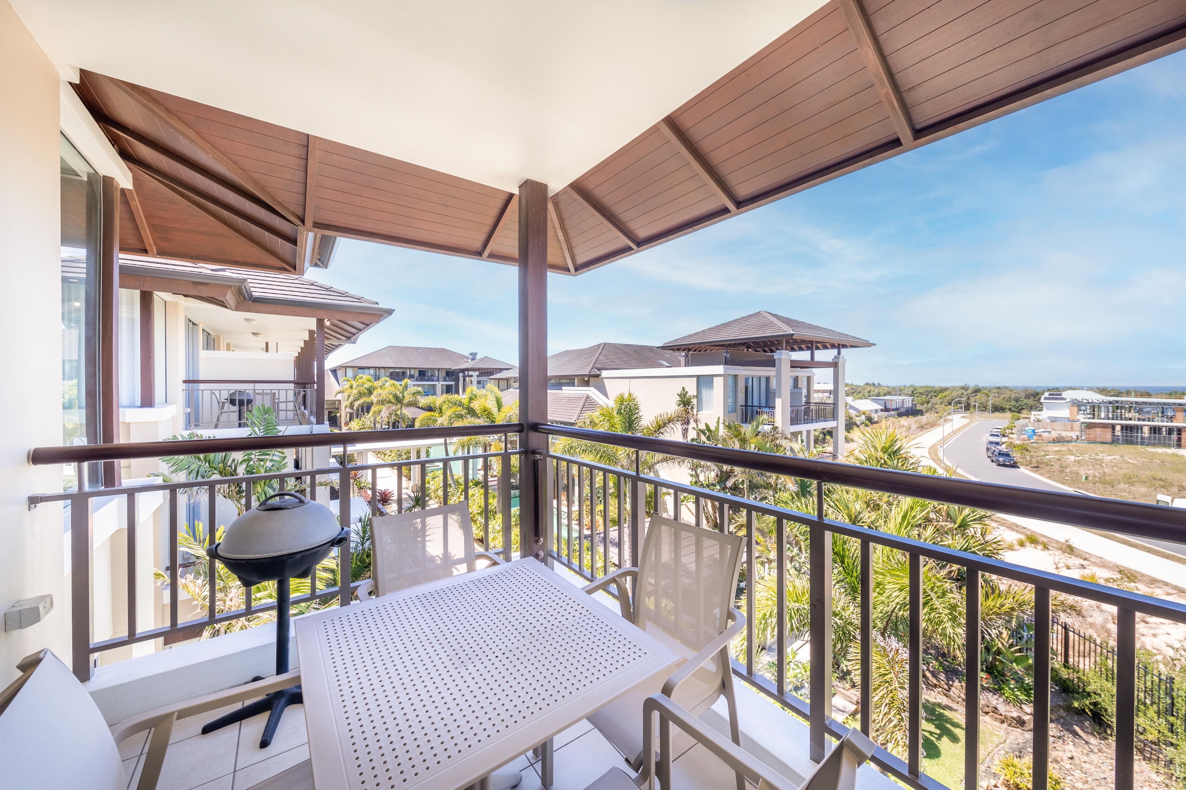 A balcony with a table and chairs, a grill, and a view of neighboring houses and a road.