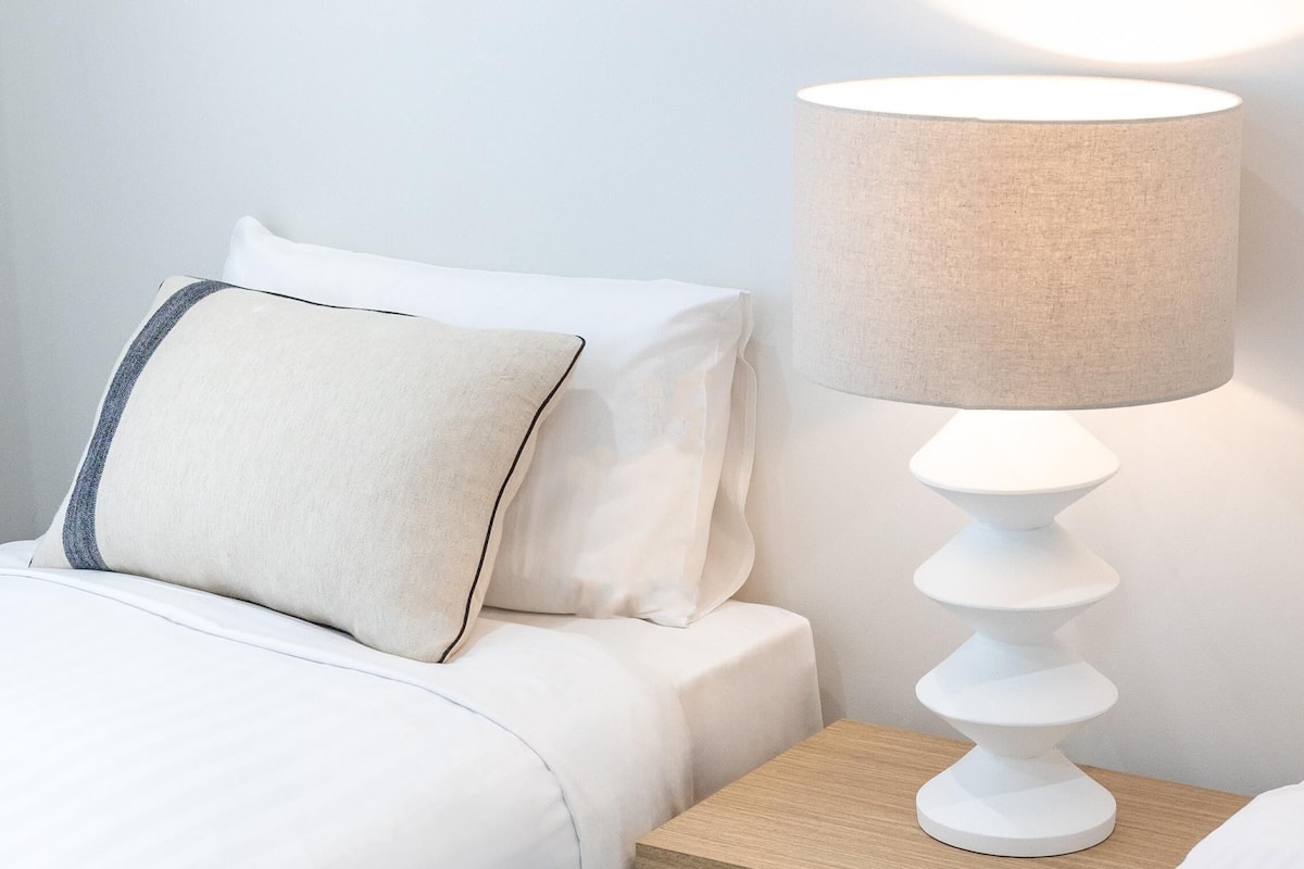 Un lit bien fait, avec un oreiller, une table de chevet sur laquelle se trouve une lampe, et un tableau encadré accroché au mur.