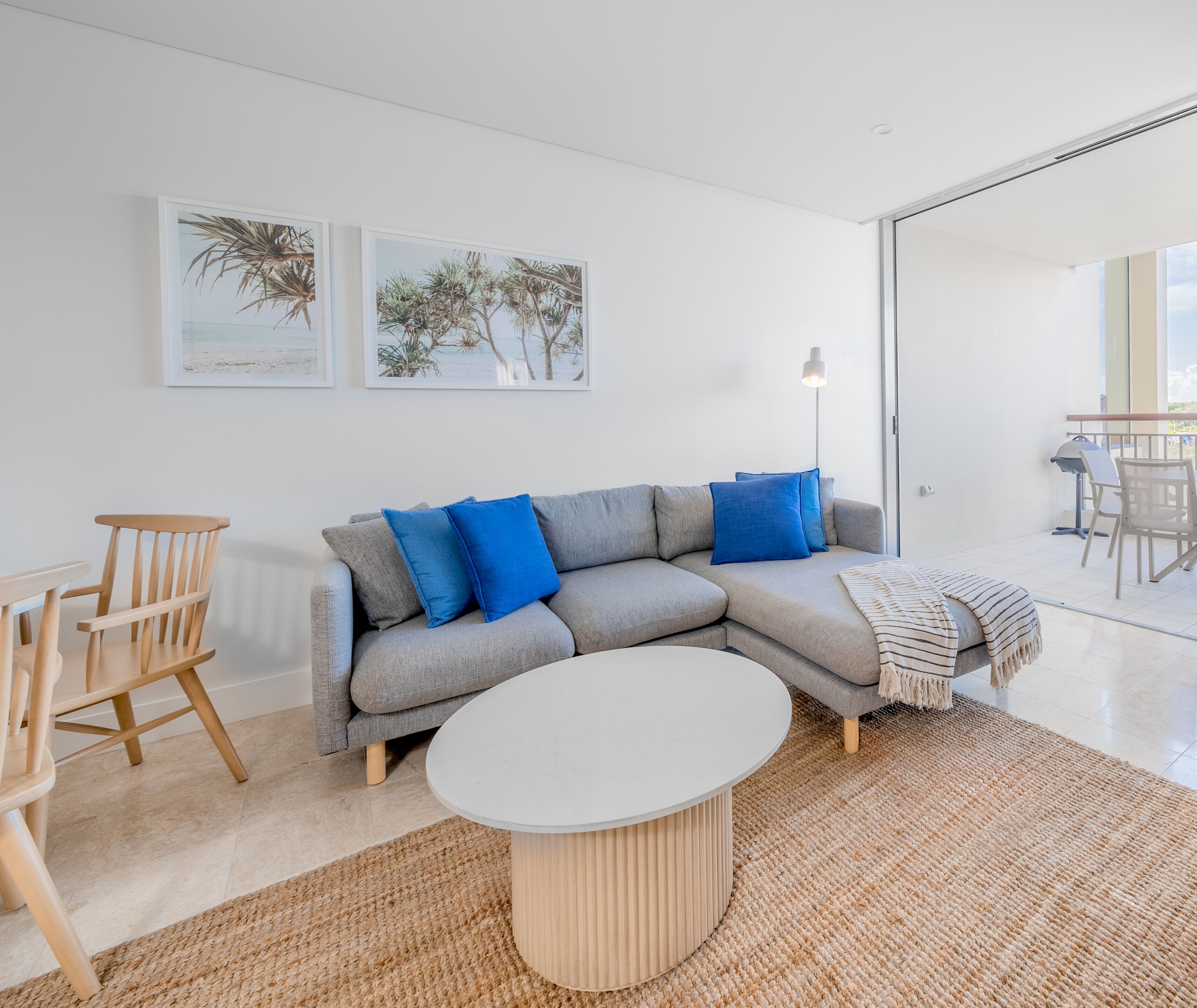 Un salon moderne avec un canapé d’angle gris, une table basse ronde et une chaise en bois. Deux tableaux encadrés ornent le mur, ainsi qu’une lampe sur pied. On aperçoit également un balcon aménagé avec du mobilier d’extérieur.