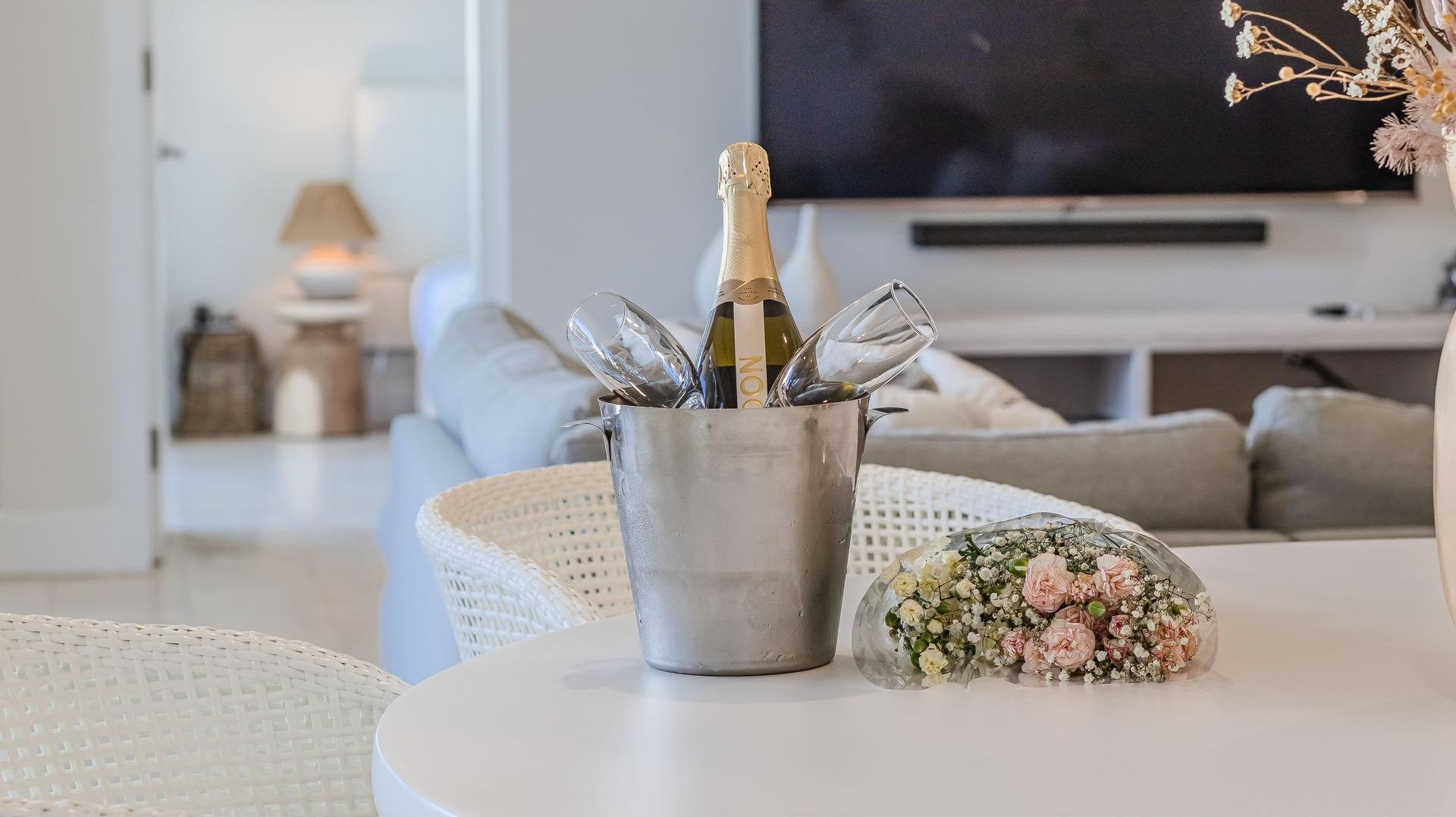 Una sala de estar con un sofá blanco, un televisor y una mesa con una botella de champán y flores.
