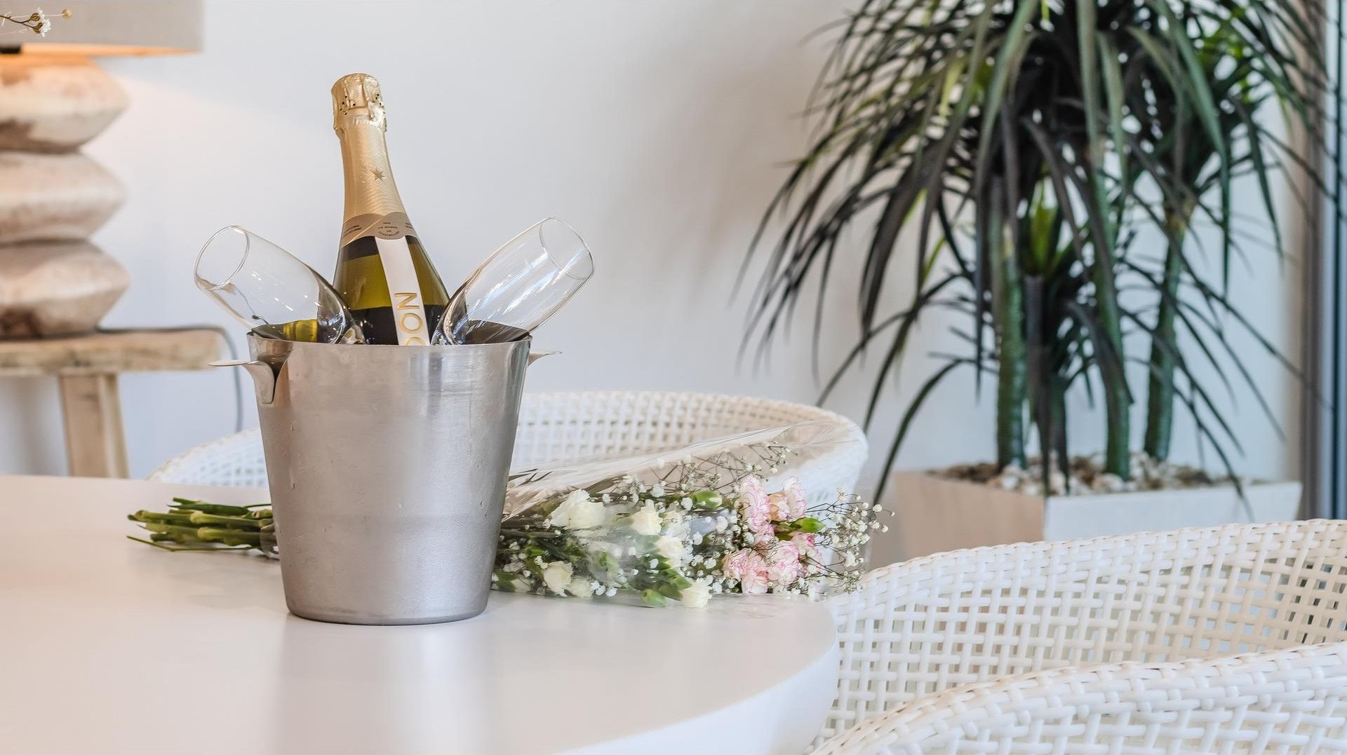 Ein Metall-Champagnerkühlbagger mit einer Flasche und Gläsern auf einem Tisch, umgeben von Korbmöbeln und einer Topfpflanze.