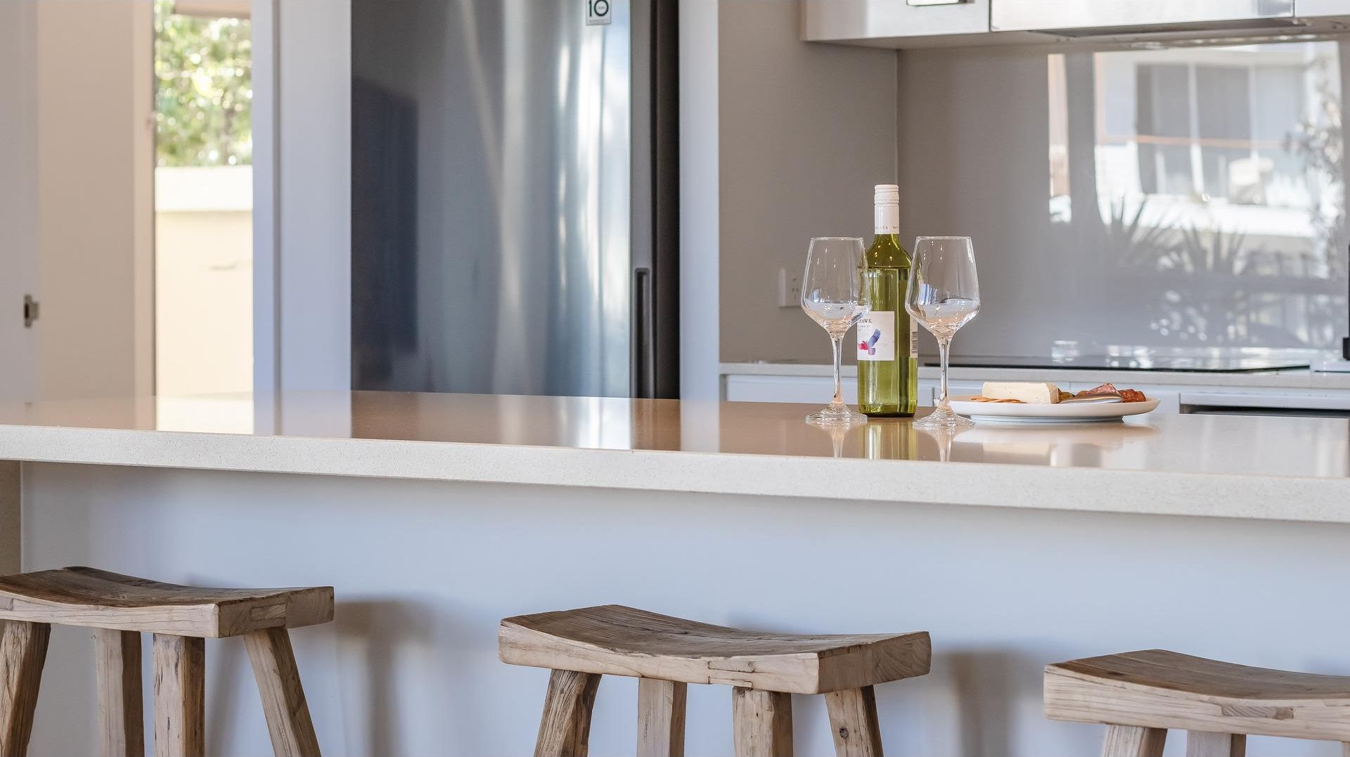 A kitchen with a stainless steel refrigerator, a wine bottle, and glasses on the counter. There are wooden stools in front.