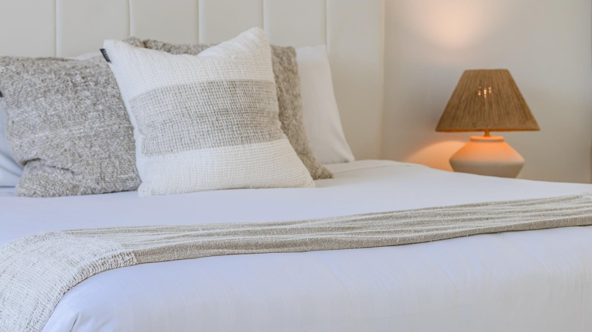 A neatly made bed with a white comforter and a striped throw blanket. There are two pillows, one with a textured pattern and the other plain. A lamp with a beige shade is on a bedside table.