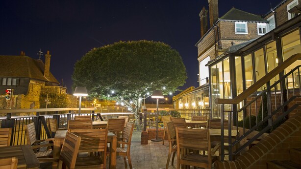 An outdoor seating area with wooden tables and chairs, a large tree, and a building with a brick facade in the background. - The Bell Hotel (Sandwich)