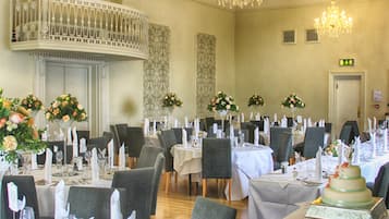 Un salón de banquetes con varias mesas redondas dispuestas para comer, cada una con manteles blancos y centros de mesa florales.