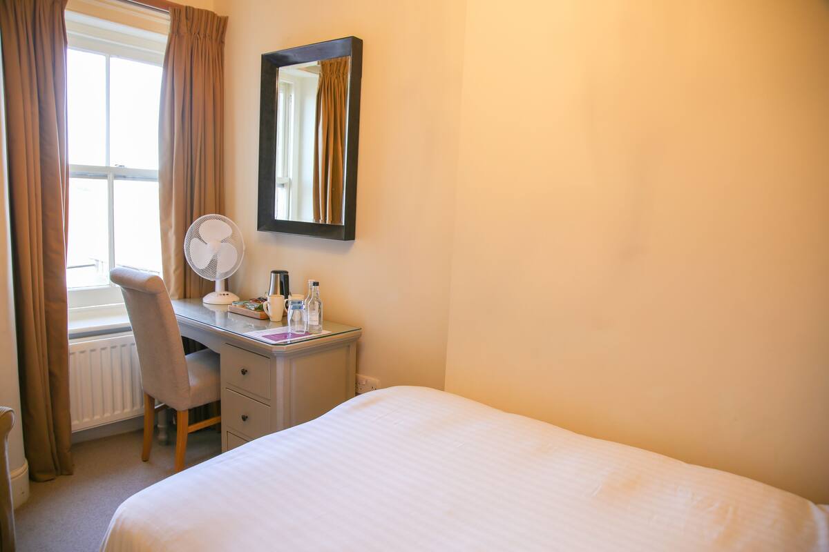 A hotel room with a bed, a desk with a chair, a mirror, a radiator, and a window with curtains.