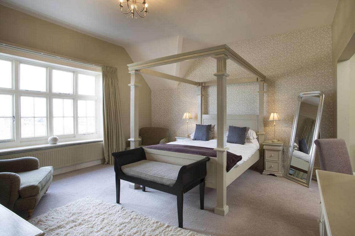 A four-poster bed, a bench, a chair, a bedside table, a dresser, a mirror, and a window with curtains.