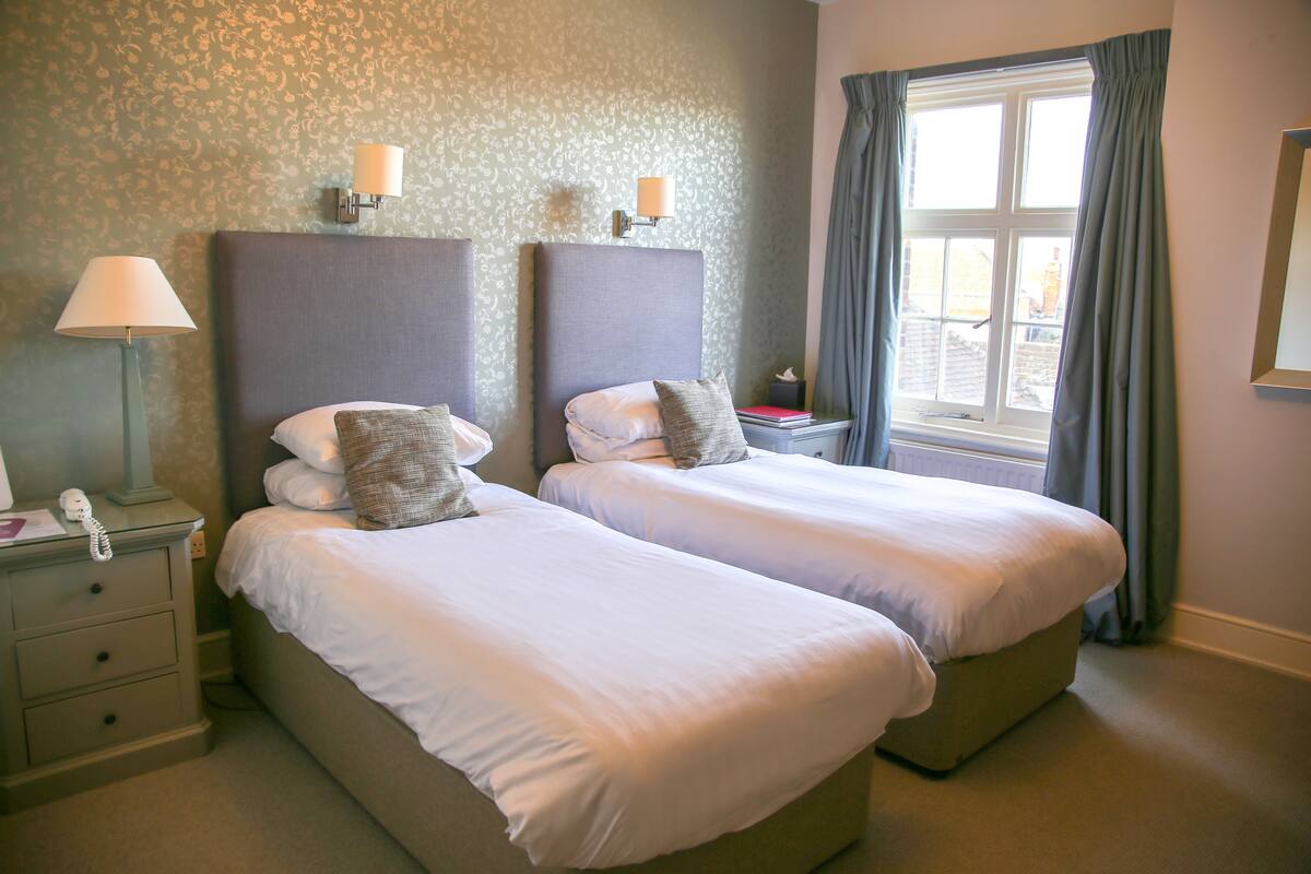 A hotel room with two beds, a nightstand with a lamp, a telephone, and a window with curtains.