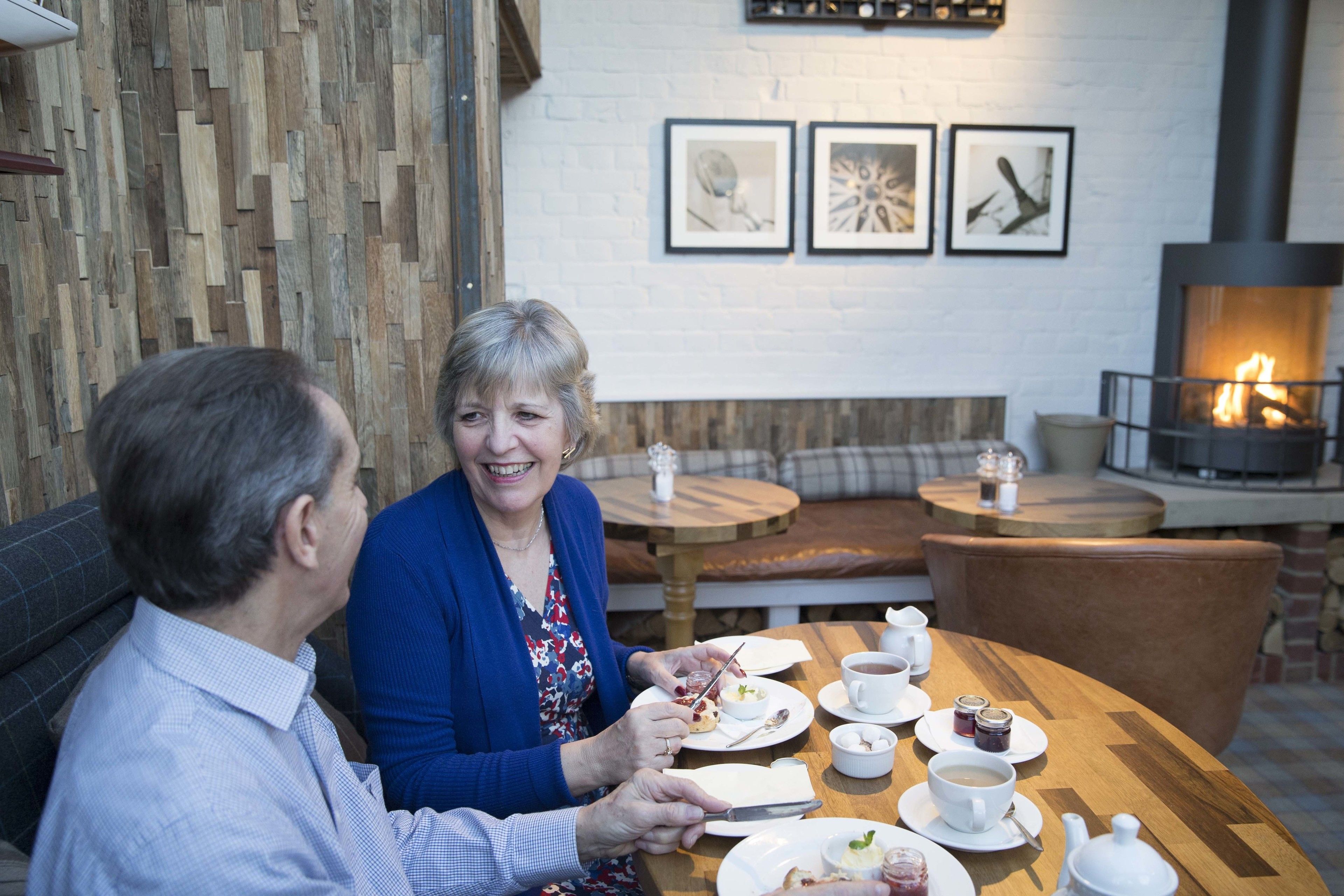 Two people dining at a wooden table with white cups, plates, and a teapot. There is a fireplace in the background and a wall with framed pictures.