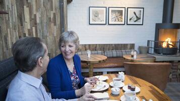 Two people dining at a wooden table with white cups, plates, and a teapot. There is a fireplace in the background and a wall with framed pictures.
