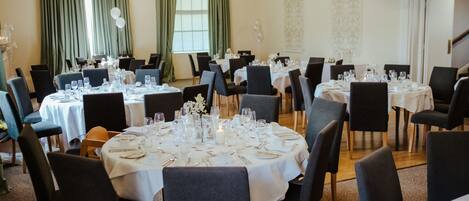A formal dining room with multiple round tables set for a meal, each with white tablecloths and dark chairs.