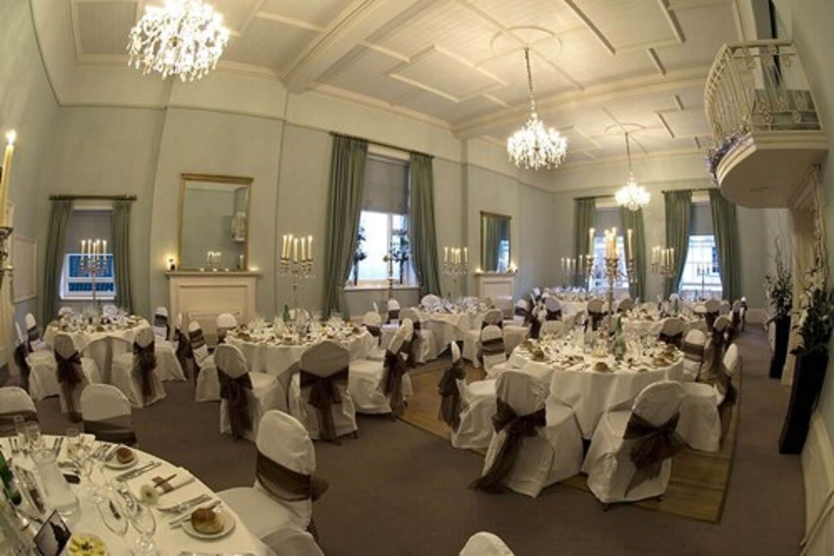 Un salón de banquetes con mesas redondas dispuestas para un evento formal, candelabros y un balcón con vistas al salón.