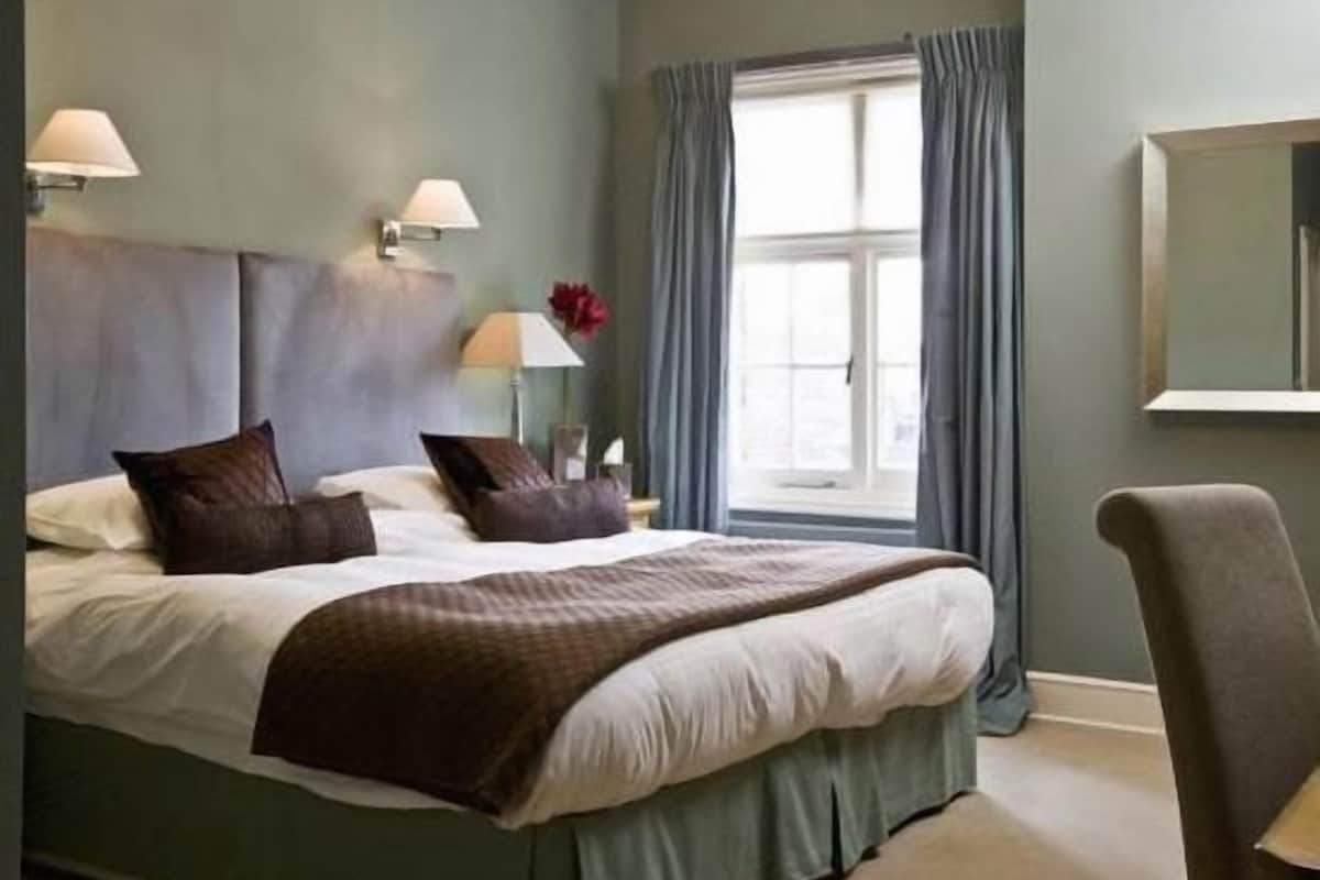 A hotel room with a large bed, a window with curtains, a chair, and a bedside table with a lamp.