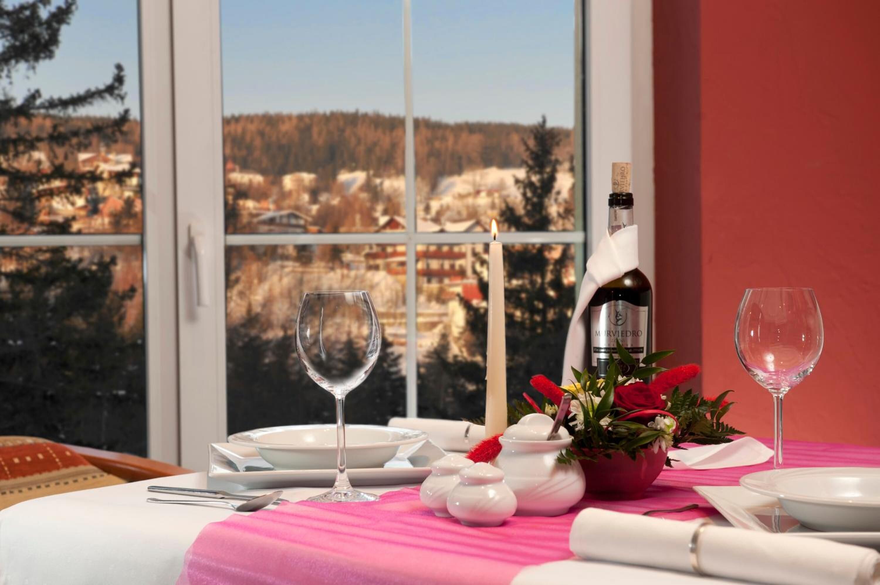 A dining table set with white plates, glasses, and a bottle of wine, with a view of a snowy landscape through a window.