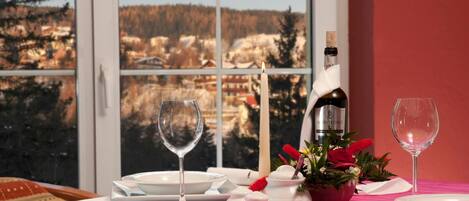 A dining table set with white plates, glasses, and a bottle of wine, with a view of a snowy landscape through a window.