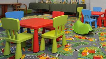 Un salón infantil con sillas y mesas coloridas, una alfombra con diseño de paisaje urbano y una mesa con libros y juguetes al fondo.