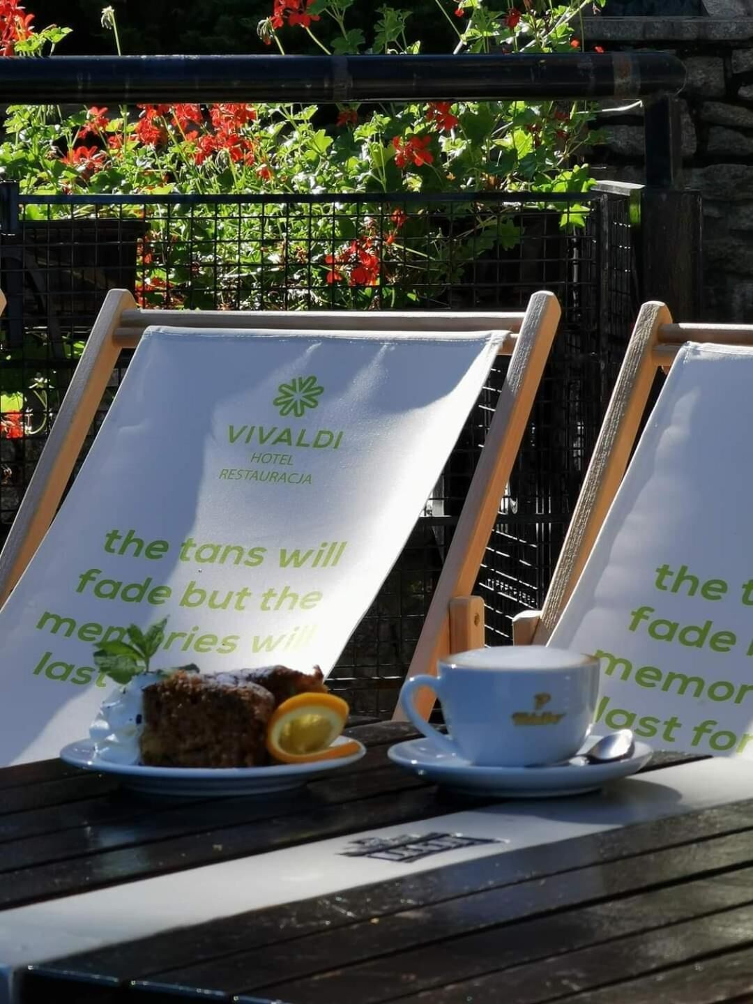 A table with a sign for Vivaldi Hotel Restaurantecia, a plate with food and a cup, and a backdrop of greenery and flowers.
