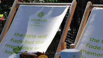 A table with a sign for Vivaldi Hotel Restaurantecia, a plate with food and a cup, and a backdrop of greenery and flowers.