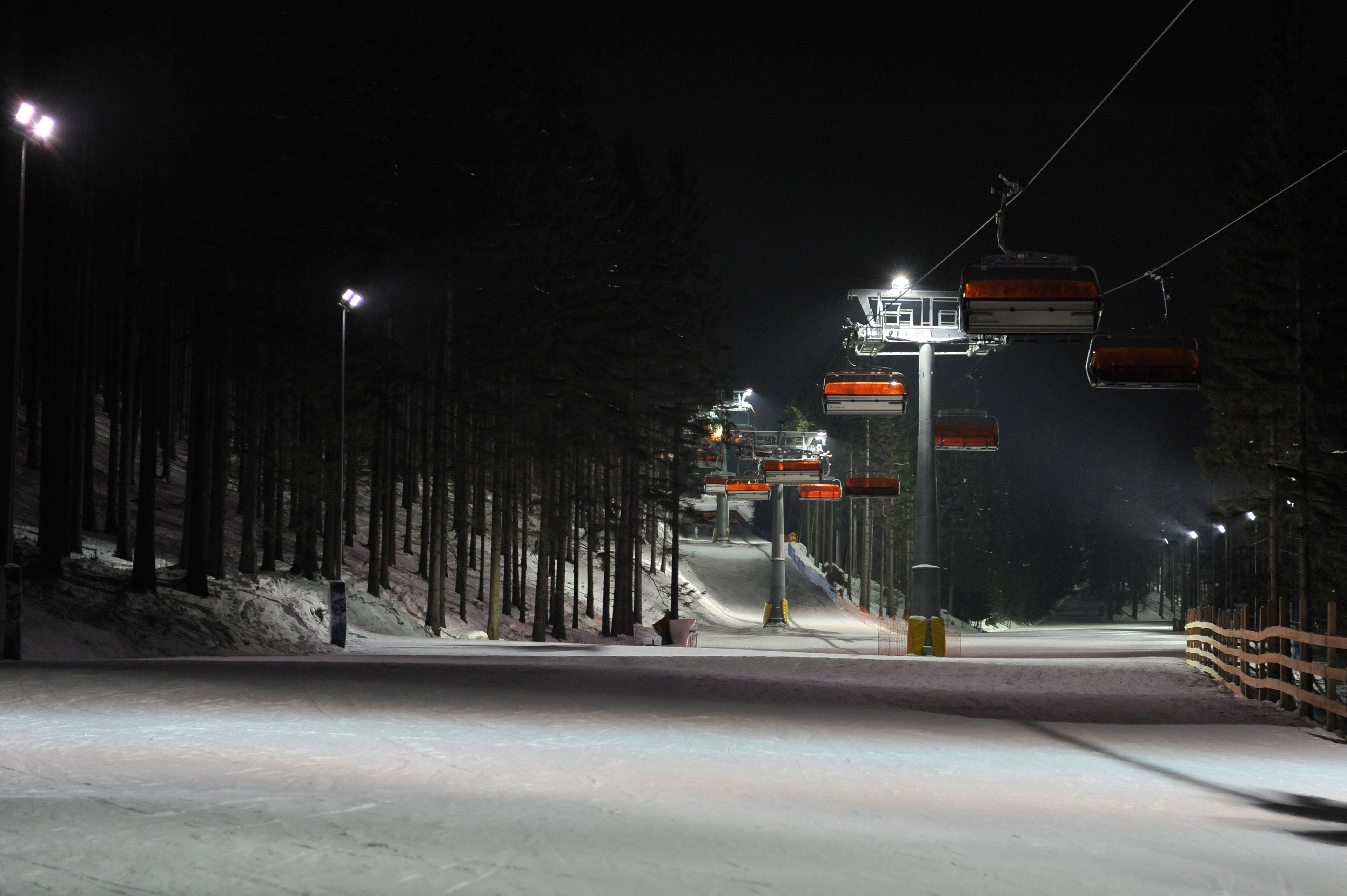 A snow-covered ski slope at night with illuminated ski lifts and a wooden fence along the side.