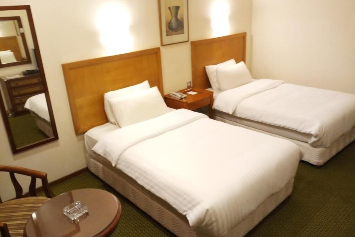 Habitación de hotel con dos camas individuales, cabecero de madera, mesita de noche con lámpara, una pequeña mesa redonda y un espejo en la pared.