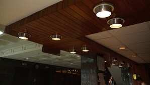 Um hall de hotel com painéis de madeira no teto, iluminação embutida e uma coluna central.
