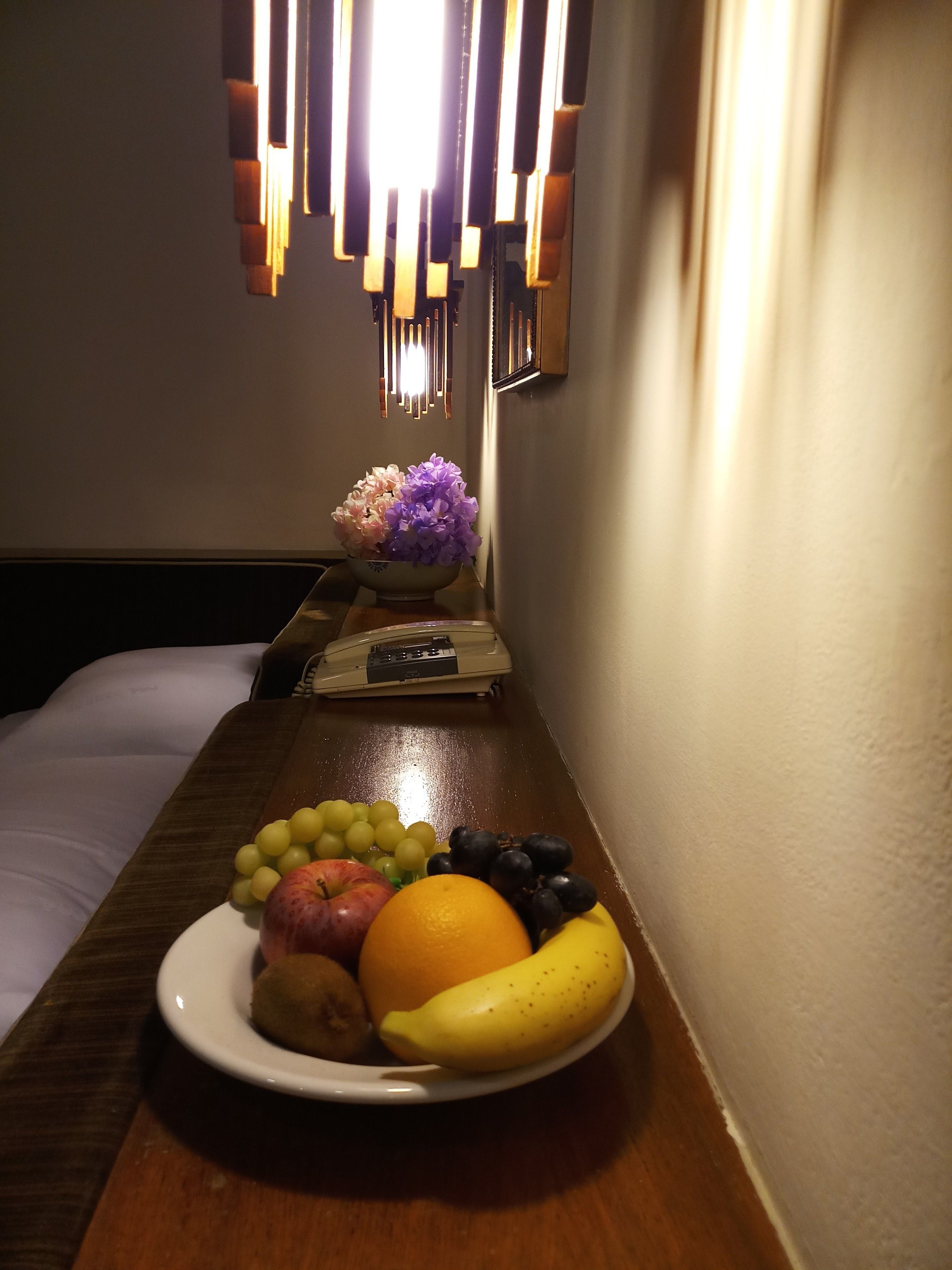 A hotel room with a wooden table displaying a plate of fruit, a vase with flowers, and a bedside table with a lamp and a remote control.