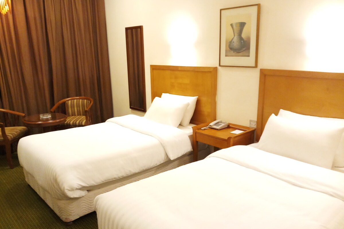 Habitación de hotel con dos camas, una mesita de noche de madera con un teléfono, una silla y un cuadro en la pared.