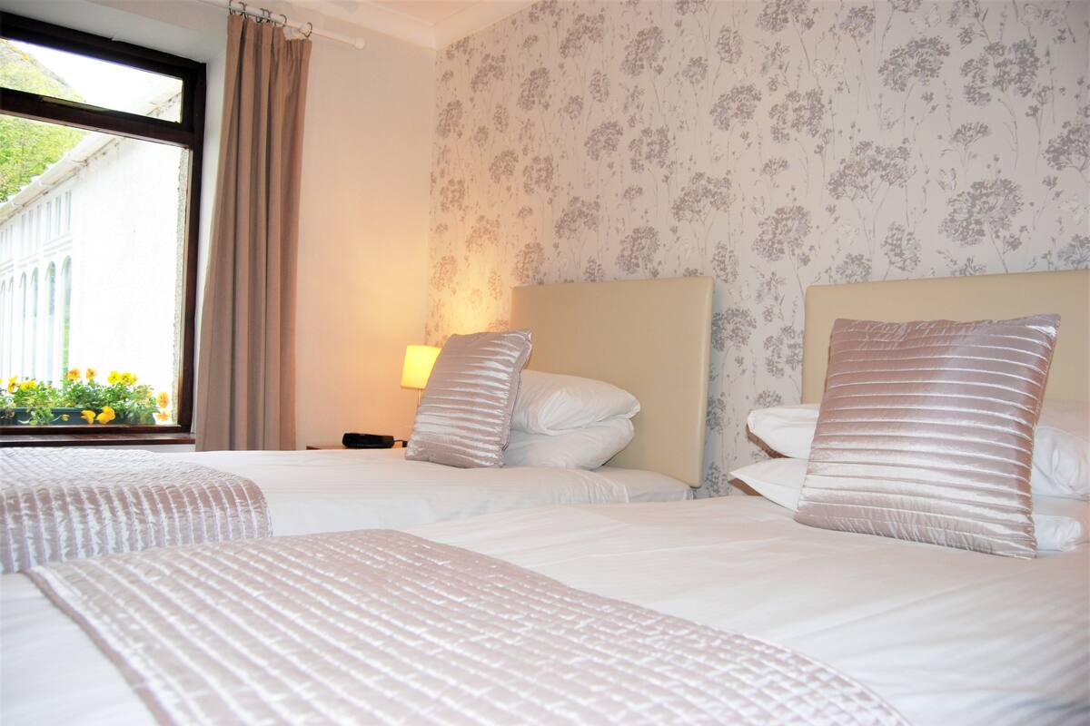 A hotel room with two beds, a window with curtains, floral wallpaper, and a nightstand with a lamp.