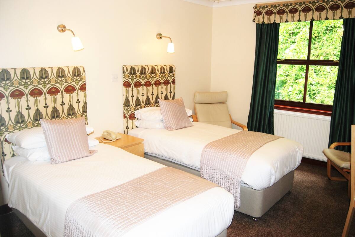 A hotel room with two beds, a chair, a nightstand, and a window with curtains.
