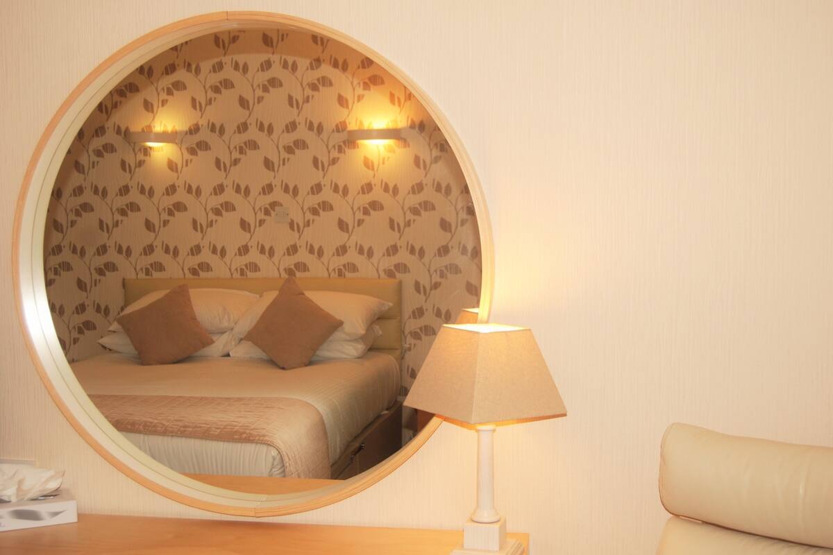 A round mirror reflecting a bed with a headboard, a bedside lamp, and a wall with a leaf pattern.