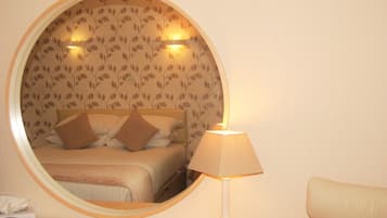 A round mirror reflecting a bed with a headboard, a bedside lamp, and a wall with a leaf pattern.