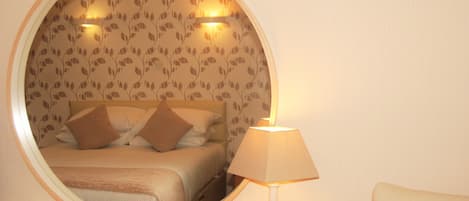 A round mirror reflecting a bed with a headboard, a bedside lamp, and a wall with a leaf pattern.