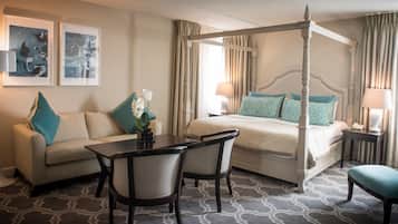 A hotel room with a four-poster bed, a sofa, a dining table, and chairs. There are two framed artworks on the wall.