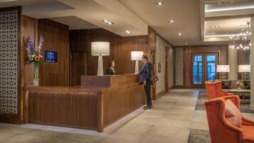 Zona de recepción de hotel con mostrador de madera, dos lámparas de pie y una pantalla digital.