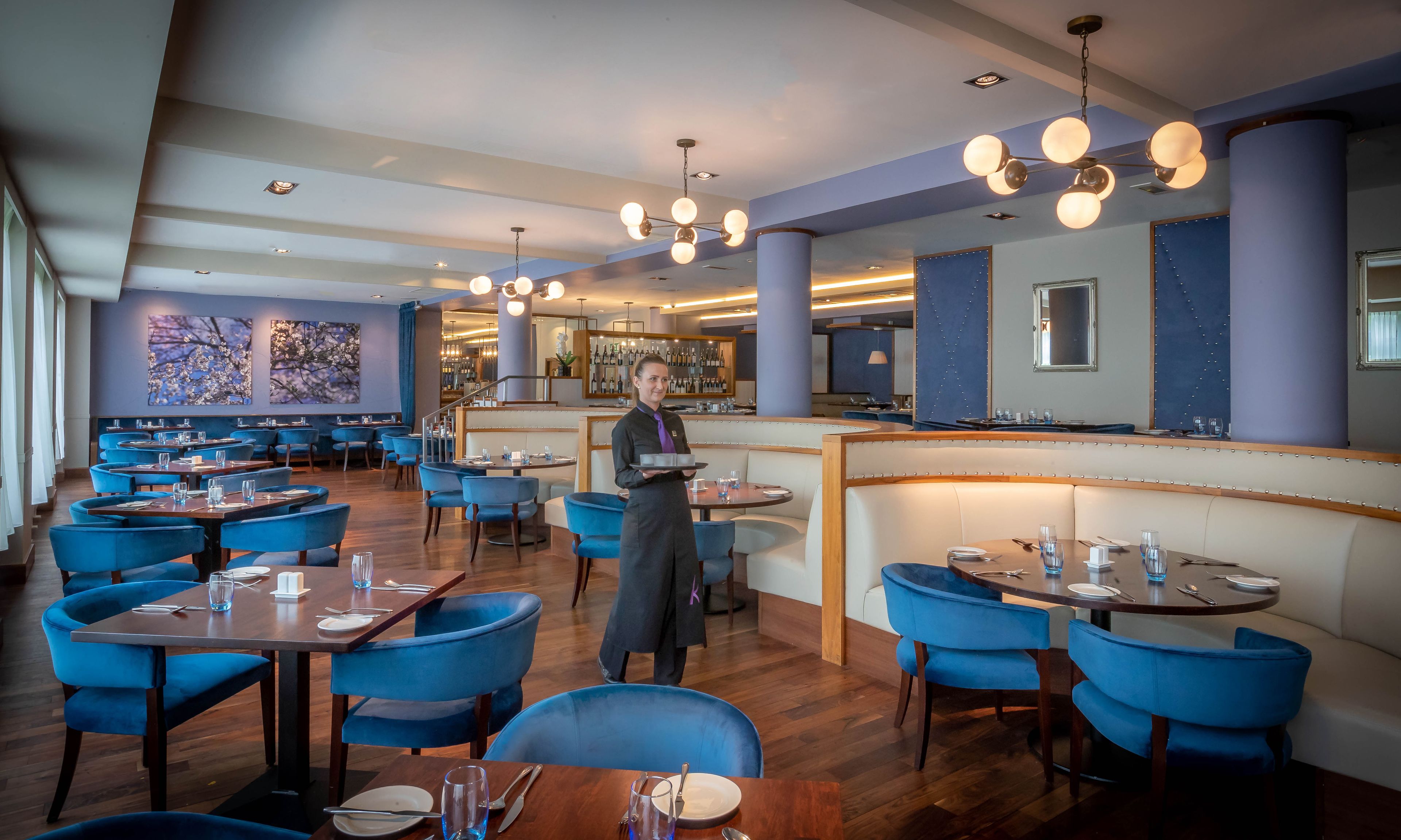 Un restaurante con sillas tapizadas en azul, mesas de madera y una zona de bar.