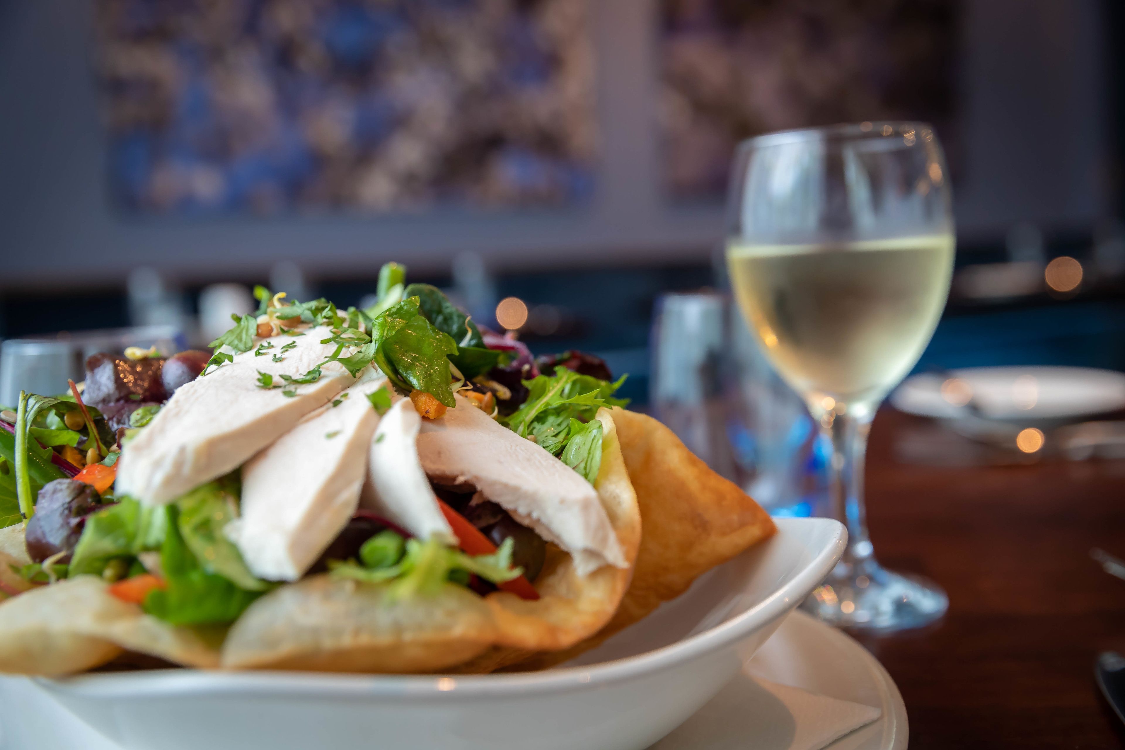 Un tazón de ensalada con pollo y verduras de hoja, una copa de vino blanco y una mesa de comedor.