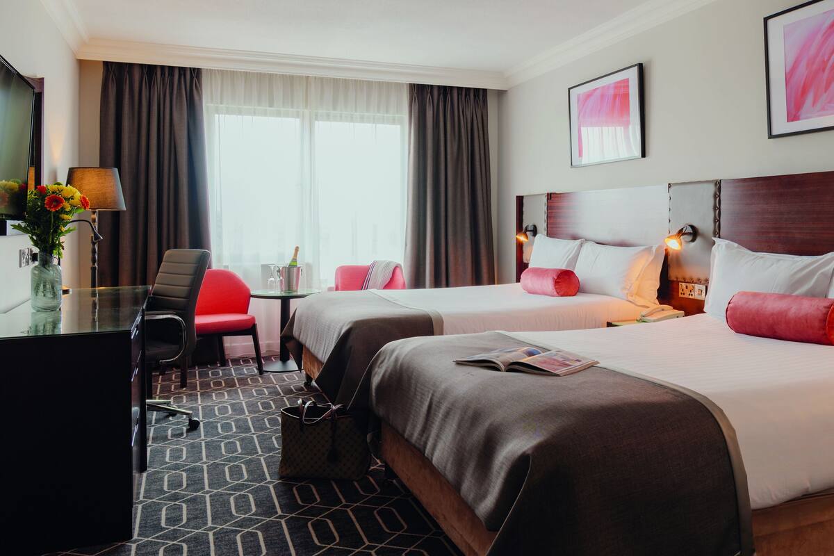 A hotel room with two beds, a desk, a chair, a vase with flowers, and a window with curtains.