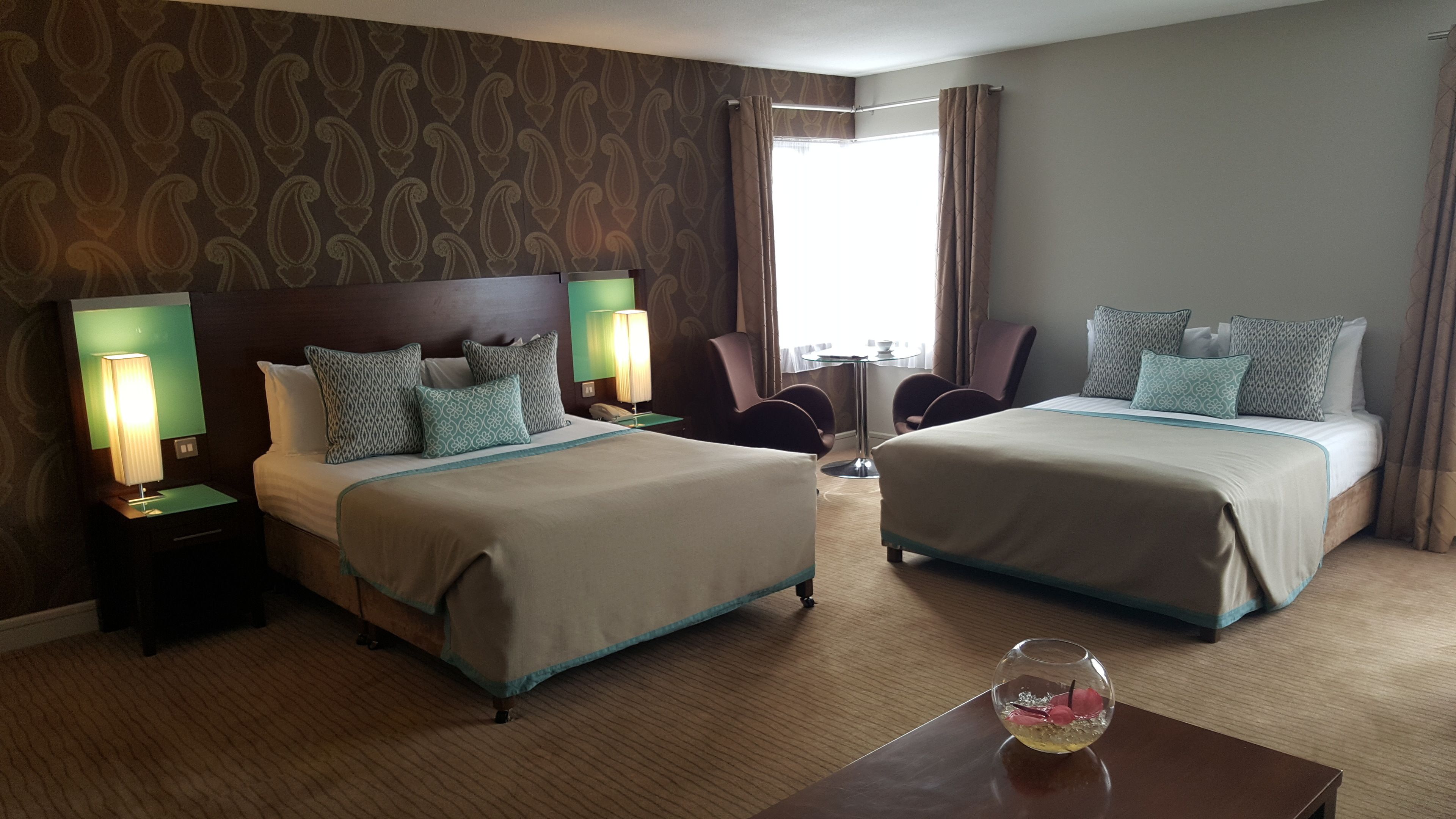 A hotel room with two beds, a nightstand with a lamp, a chair, a small table with a vase, and a window with curtains.