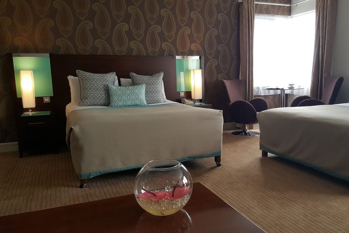 A hotel room with two beds, a fishbowl on a table, and a view of the outdoors.