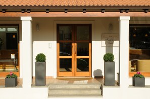 L’entrée d’un hôtel, avec une porte à double battant en bois, deux plantes en pot et une enseigne indiquant « Hôtel Angedras ».
