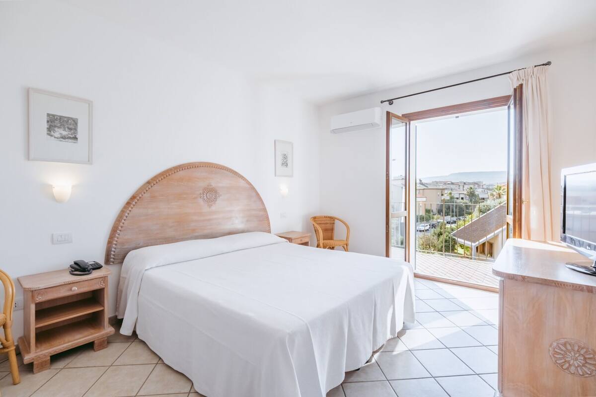 A hotel room with a large bed, a wooden headboard, a desk with a chair, a television, and a balcony with a city view.